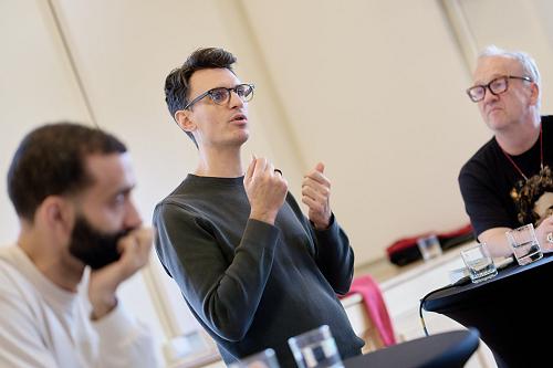Un homme parle dans ce qui semble être une conférence, entouré de deux autres qui écoutent