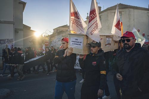 Des hommes portent sur leurs épaules u cercueil sur lequel il est apposé des feuilles sur lesquelles il est écrit « FNE fossoyeur de la ruralité ». Du cercueil s'élèvent aussi des drapeaux blanc et rouge des Jeunes agriculteurs. 