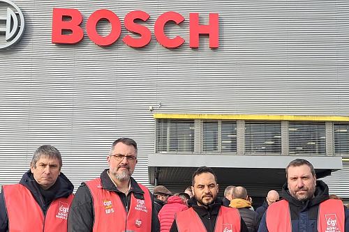 Quatre hommes en chasubles rouge de la CGT, debout devant l'entrée d'une usine sur laquelle est écrit Bosch en lettres rouges, regardent vers l'objectif.