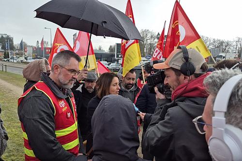 Une femme parle avec des hommes en chasubles rouge et avec des drapeaux de la CGT, un homme filme. 