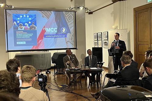 Trois hommes, dont un avec un micro, devant une salle d'une soixantaine de personnes