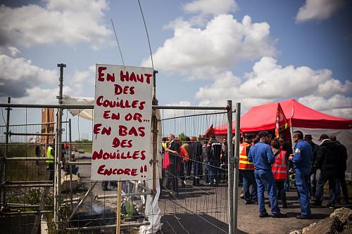 une affiche "en haut des couilles en or, en bas des nouilles encore" devant un barnum et des hommes en bleu de travail et gilets à haute visibilité