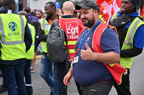 Un homme portant un chasuble rouge, souriant et levant le pouce, passe devant un groupe d'hommes portant des chasubles VGT et des chasubles Geodis.