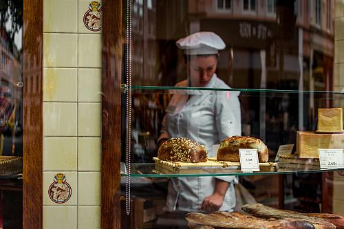 une femme en toque et tablier blancs derrière les vitres d'une boulangeries regarde les étals. 