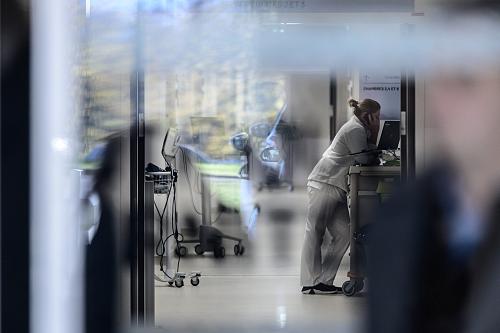 Une femme en uniforme blanc fatiguée au téléphone derrière une porte vitrée avec des reflets dans un couloir d'hôpital.