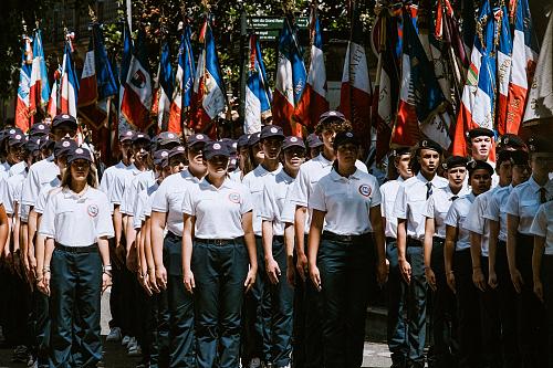 des jeunes, casquettes bleues, tee-shirts blancs, pantalon bleu, en rang et au garde-à-vous devant une rangée de drapeaux français