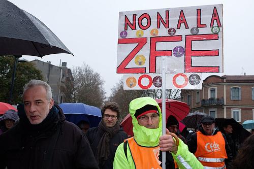 Dans une manifestation, un homme qui porte un chasuble CFDT tient une pancarte sur laquelle il est écrit « Non à la ZFE » et sur laquelle des vignette de Crit'Air sont collées.