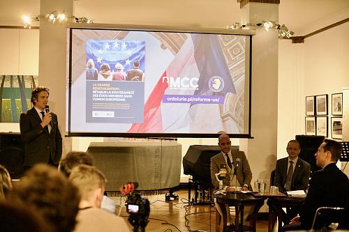 Trois hommes, dont un avec un micro, devant une salle d'une soixantaine de personnes