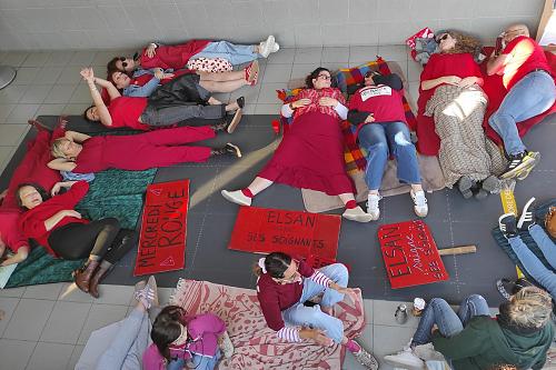 Des personnes vêtues de rouges sont allongées par terre à côtés de pancartes rouges. Sur l'une d'elle on peut lire "Elsan saigne ses soignantes".