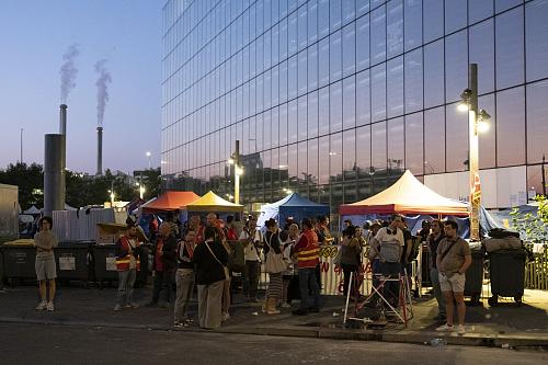 Un groupe de personnes, debout, de nuit, regroupées autour de lampadaires et de quelques stands. Certaines portent de chasubles rouge. 