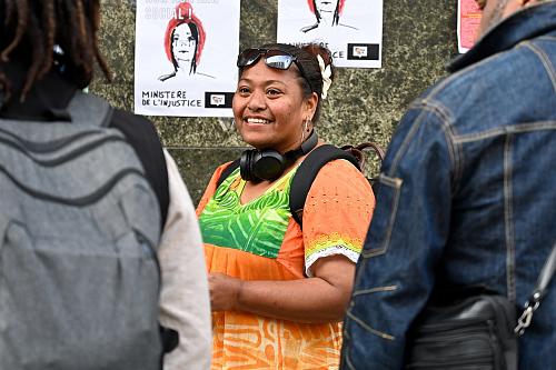  Brenda Wanabo-Ipeze, habillée en orange, sourit, devant une affiche sur laquelle il ets écrit « ministère de l'injustice ». Elle regarde evrs deux personnes qui sont dos à l'objectif. 