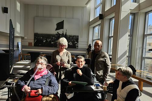 Un groupe de 5 personne se tient dans un espace lumineux. Deux personnes sont assises en fauteuil roulant, d'autres sont debout autour d'elles. En arrière-plan, de grandes fenêtres et une photographie historique accrochée au mur.