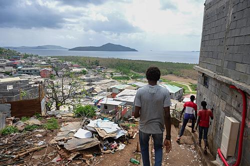 Trois jeunes hommes vus de dos regardent un paysage de bidonville de maisons de tôles depuis le haut d'une colline. 