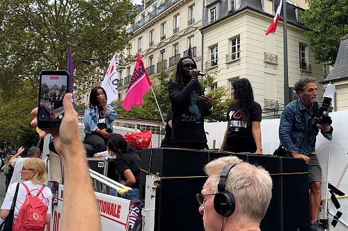Plusieurs personnes sont à l'arrière d'un camion. A l'avant un homme parle dans un micro. Au premier plan, une personne filme avec son téléphone.