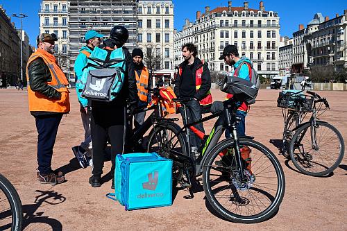 Des livreurs a vélos Uber Eats et Deliveroo portant des gilets CGT et Union indépendants discutent ensemble à côté de vélos et de sacs de livraison. 