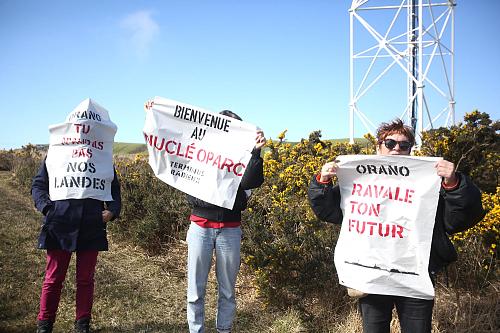  Trois personnes au bord d'une route porte des affiches dans le vent : « Orano , tu n'auras pas nos landes », « Bienvenu au nucléoparc », et « Orano, ravale ton futur ». 