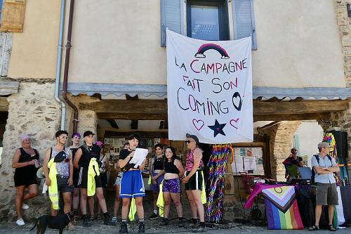 Une dizaine de personnes devout devant un bâtiment, une personne lit un texte au micro, une banderole sur laquelle il est écrit "La campagne fait son coming out" est accrochée au bâtiment.