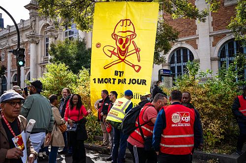 Des manifestants dont certains avec un chasuble rouge de la CGT devant une banderole jaune sur laquelle est dessinée en rouge un tête de mort coiffée d'un casque de chantier et est écrit « BTP, un mort par jour ». 