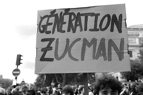 Un homme dans une manifestation tient une pancarte « Génération Zucman », la photo est en noir et blanc. 
