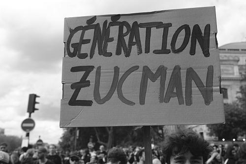 Un homme dans une manifestation tient une pancarte « Génération Zucman », la photo est en noir et blanc. 