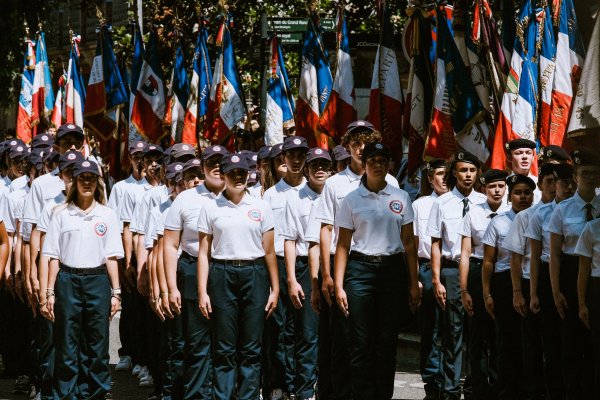 des jeunes, casquettes bleues, tee-shirts blancs, pantalon bleu, en rang et au garde-à-vous devant une rangée de drapeaux français