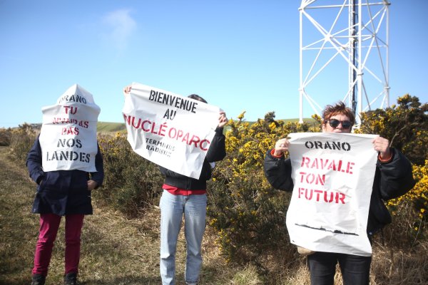  Trois personnes au bord d'une route porte des affiches dans le vent : « Orano , tu n'auras pas nos landes », « Bienvenu au nucléoparc », et « Orano, ravale ton futur ». 