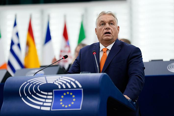 Viktor Orbán parle à un pupitre sur lequel on voit le symbole d'un hémicycle et le drapeau européen. On voit des drapeaux au deuxième plan. 