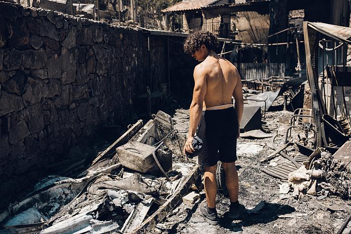 Un jeune homme en short et torse nu regarde au sol les cendres d'objects calcinés dans ce qu'il reste d'une cour ou d'une ruelle.