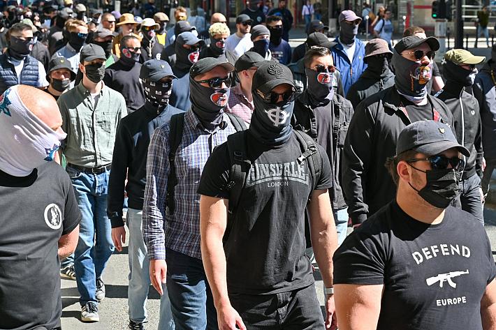Militants d'extrême droite défilant en rang, masqués, arborant des croix celtiques ou des tee-shirts "Defend Europe" avec un fusil d'assaut.