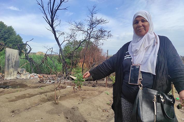 Une femme portant un foulard, un téléphone portable à son cou et un sac à main montre un terrain en friche, brûlé par les flammes.