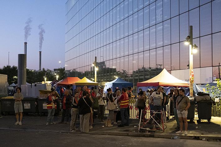 Un groupe de personnes, debout, de nuit, regroupées autour de lampadaires et de quelques stands. Certaines portent de chasubles rouge. 