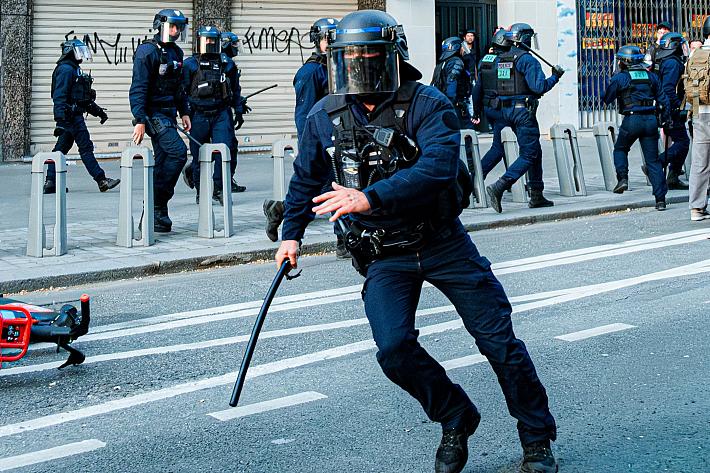 Un policier casqué court avec une matraque à la main dans une rue, d'autres policiers en uniforme de maintient de l'ordre se trouvent au second plan.