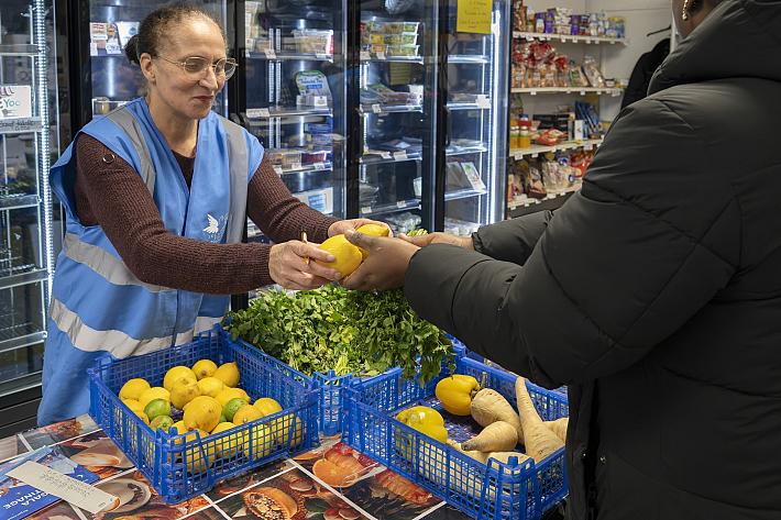 Une femme portant un gilet bleu avec le logo de l'association du Secours populaire tend des fruits et légumes a une personne pauvre venue chercher une aide alimentaire. De nombreux légumes poses sur une table sont également disponibles ainsi que des produits laitiers visibles dans un grand réfrigerateur aux portes transparentes.
