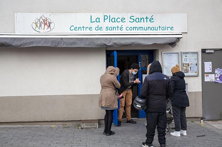 Des agenrs à l'entrée d'un bâtiemtn sur lequel il est écrit "Place Santé, centre de santé communautaire". Un homme prend des inscriptions.