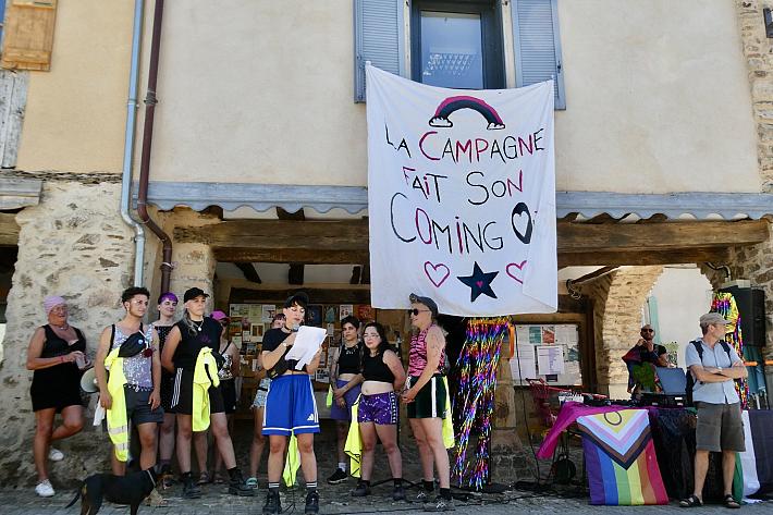 Un groupe d'une dizaine de personne devant un bâtiment, une personne lit un texte au micro, une banderole sur laquelle il est écrit "La campagne fait son coming out” est accrochée à la fenêtre du bâtiment.