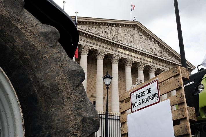 une roue de tracteur, un panneau "fiers de vous nourrir" et au fond la façade de l'assemblée nationale