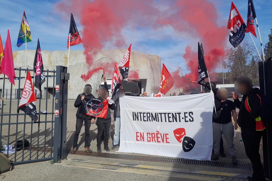 Huit personnes aux visages floutés tenant des drapeaux CNT t des fumigènes rouge debout derrière une banderole sur laquelle il est écrit « Intermittents en grève ». 