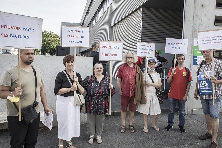 Sept personnes debout devant un bâtiment tiennent des pancartes sur lesquelles il est écrit « Pouvoir payer ses factures » ; « Je ne suis ni un fainéant, ni un mendiant » ; « Vivre, aps survivre » ; « Je veux qu'on m'aide, pas qu'on me malmène » ; « Le RSA, c'est la misère ». 
