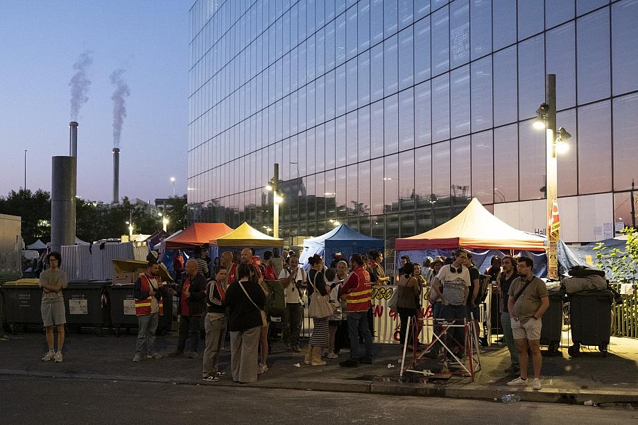 Un groupe de personnes, debout, de nuit, regroupées autour de lampadaires et de quelques stands. Certaines portent de chasubles rouge. 