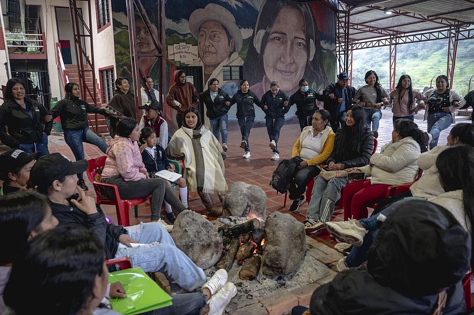 Un groupe de personne assises en rond dans un local. Des jeunes dansent derrière une danse qui semblent être traditionnelle. 
