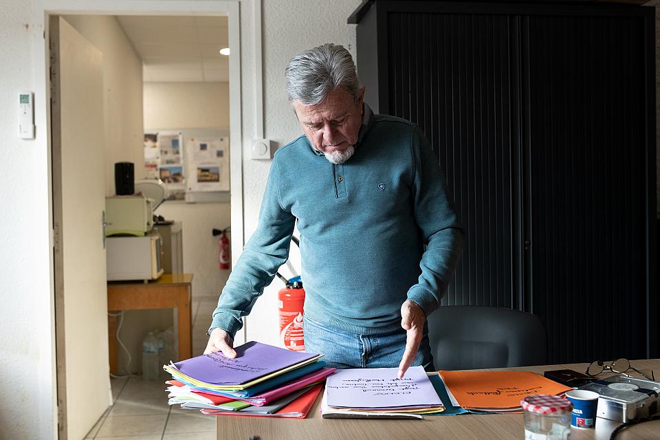 Daniel Moutet, un monsieur d'une soixantaine d'année portant un pull bleu, une barbe, avec face à lui plusieurs dossiers de couleurs différentes, dans les locaux de l'Association Défense et Protection du Littoral du Golfe de Fos, en février 2026. 