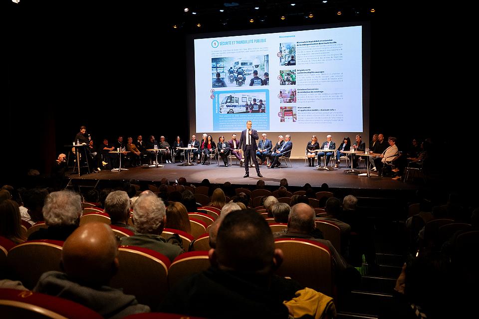 Rémi Esnault sur la scène du théâtre municipal de Fos-sur-Mer, en costard. Derrière lui tous ses colistiers, assis sur des chaises. Sur l'écran apparaît le début de son programme, sur la sécurité.