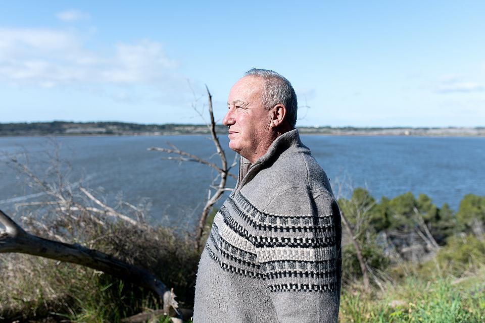 Jean-Louis Sanial, président du collectif Sauvons nos étangs, portant un pull gris, de profil, devant un grand étang de Fos-sur-Mer par beau temps.