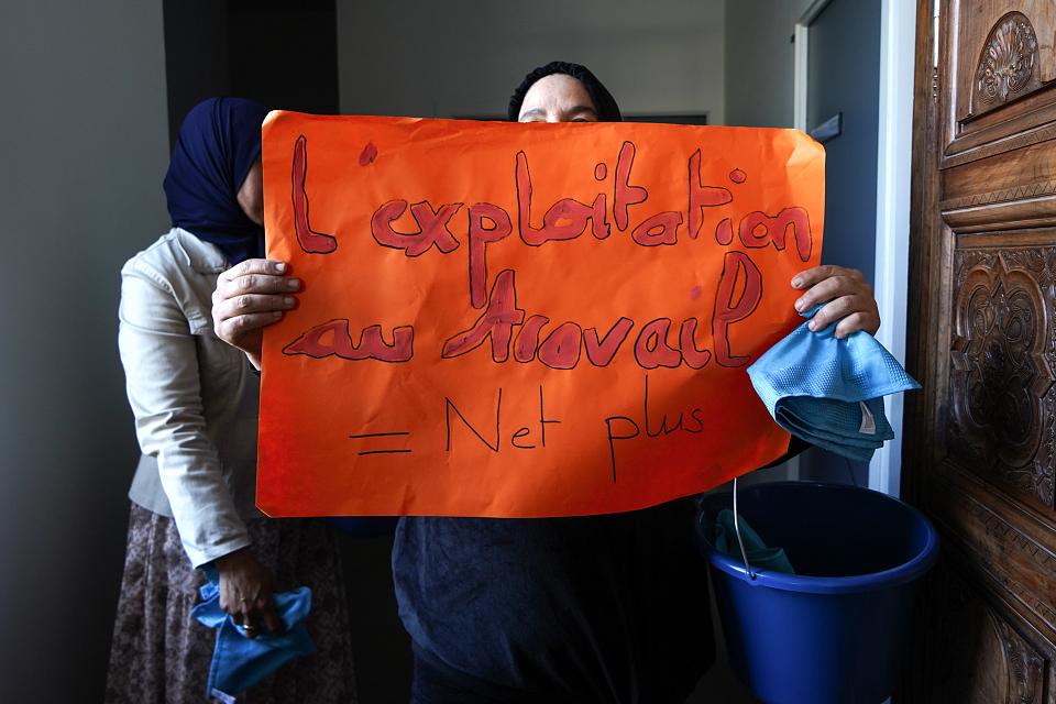 Une femme porte devant son visage une afficheorgange où il est écrit "L'exploitation au travail = Net Plus3
