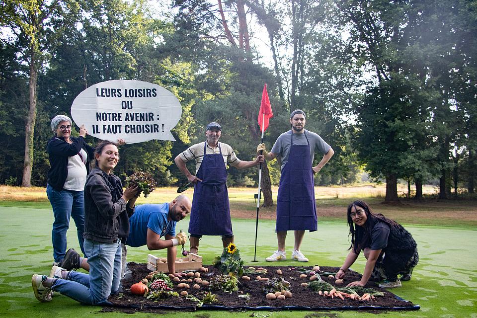 Six personnes sont rassemblées autour de légumes posés sur une bâche recouverte de terre. Deux personnes tiennent un drapeau du parcours de golf, où le potager est installé. Un message est inscrit sur une pancarte : "Leurs loisirs ou notre avenir : il faut choisir !"