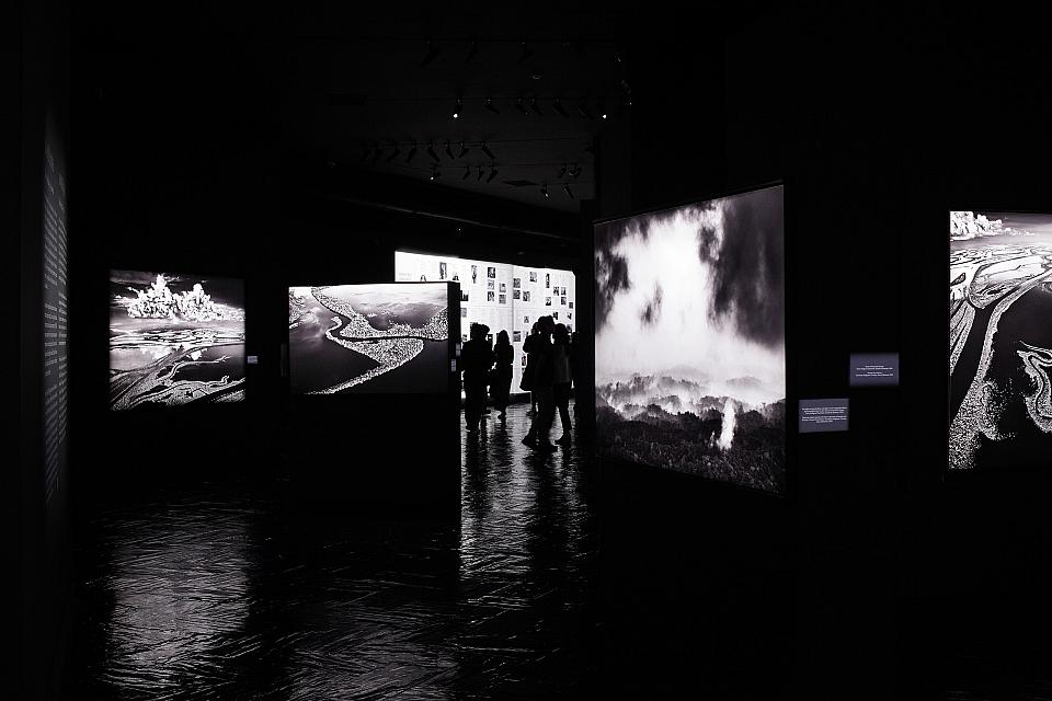 Une image en noir et blanc d'une exposition, où l'on devine des silhouettes au milieu des photos.