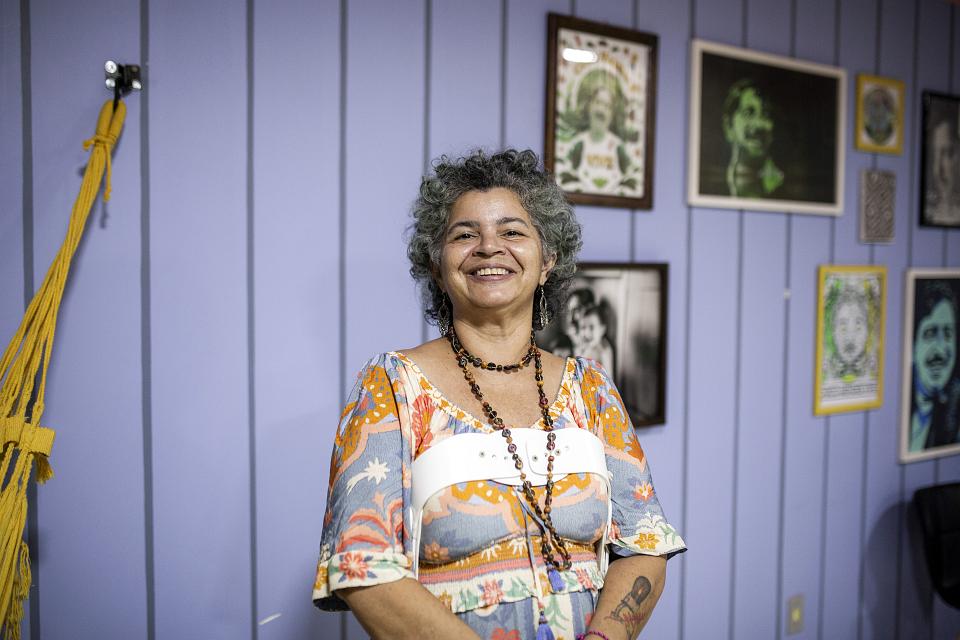 Une femme souriante devant un mur mauve sur lequel sont accrochées des photos. 