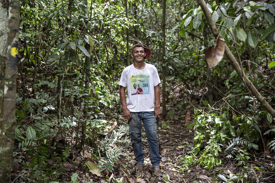 Un jeune homme souriant debout dans la forêt. Il porte une casquette rouge, un jean et un t-shirt blanc. 