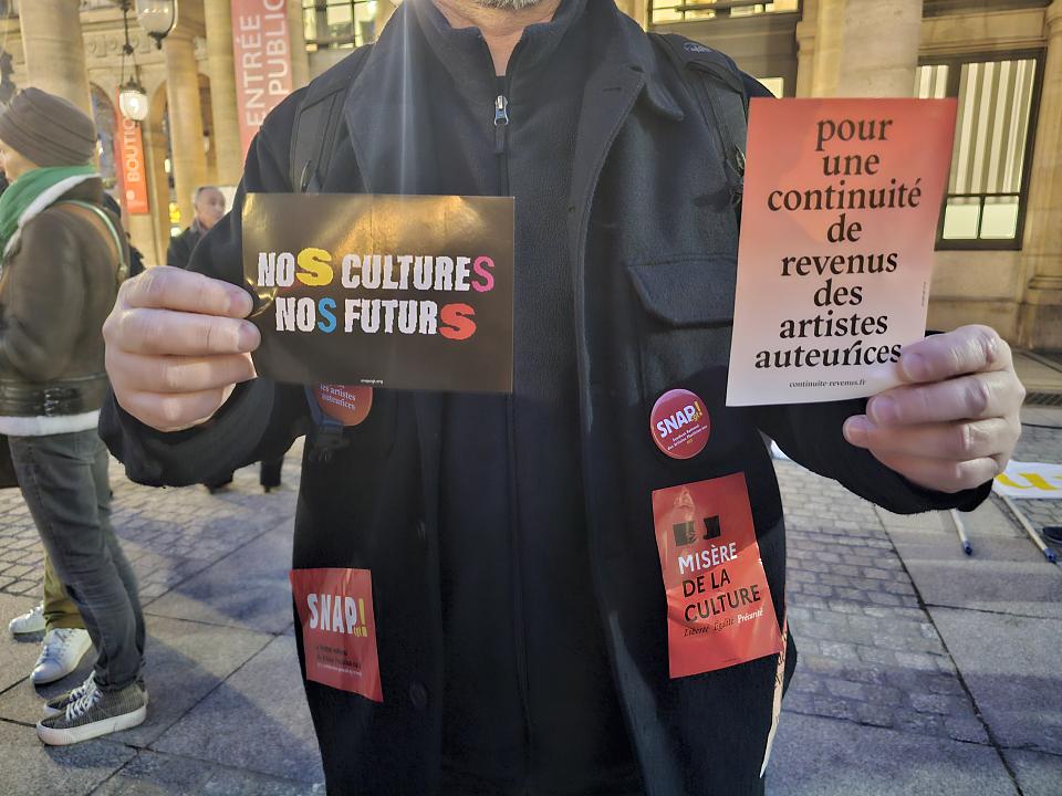 Une personne dont on en voit pas le visage tient deux affiches "Nos cultures, nos futurs" et "pour une continuité de revenus des artistes auteurices"