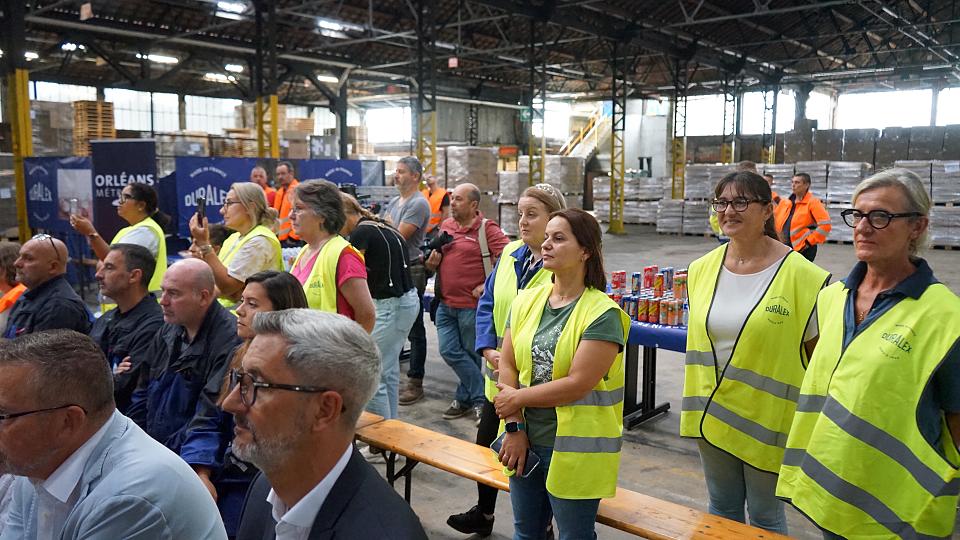 Des salariées en chasuble jaunes et d'autres personnes sur des bancs dans un hangar